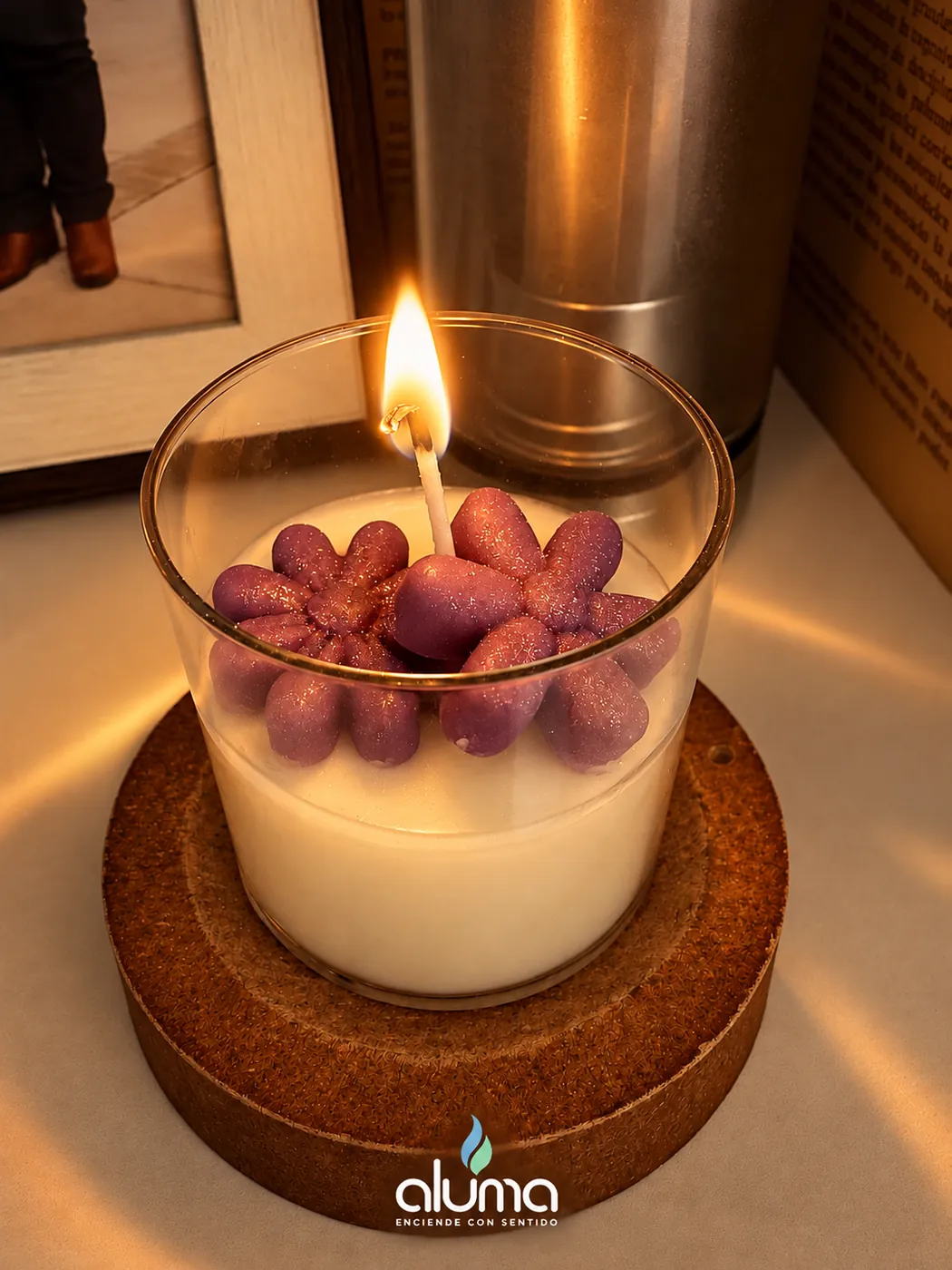 Vela en vaso de vidrio con flores de cera moradas, encendida sobre una base de madera